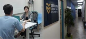 一名穿着西装外套的女性工作人员在办公室与一名穿蓝色T恤的男性面对面交流,桌上摆放着笔记本电脑和文件。旁边的墙上挂着‘UNITED LISEN Education Centre 联邦乐尚雅思中心’的标识,右侧是教育中心的走廊,墙壁深蓝色,摆放着绿植。